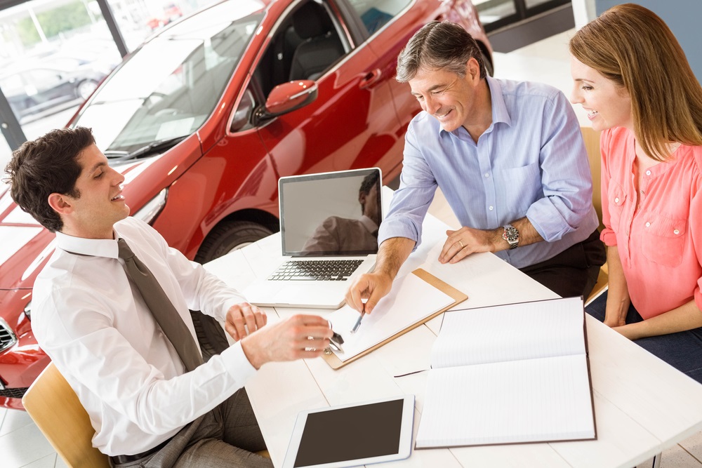 Employee speaking with two customers across a desk covered with car finance paperwork inside a car dealership