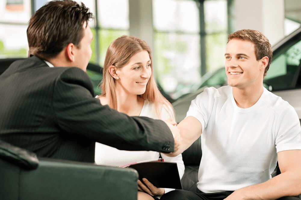 Employee shaking hands with one customer, sitting next to another customer inside a car dealership