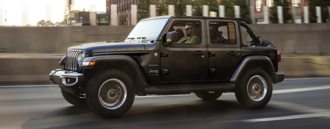 A grey 2022 Jeep Wrangler is shown from the side driving on an open road through a city.