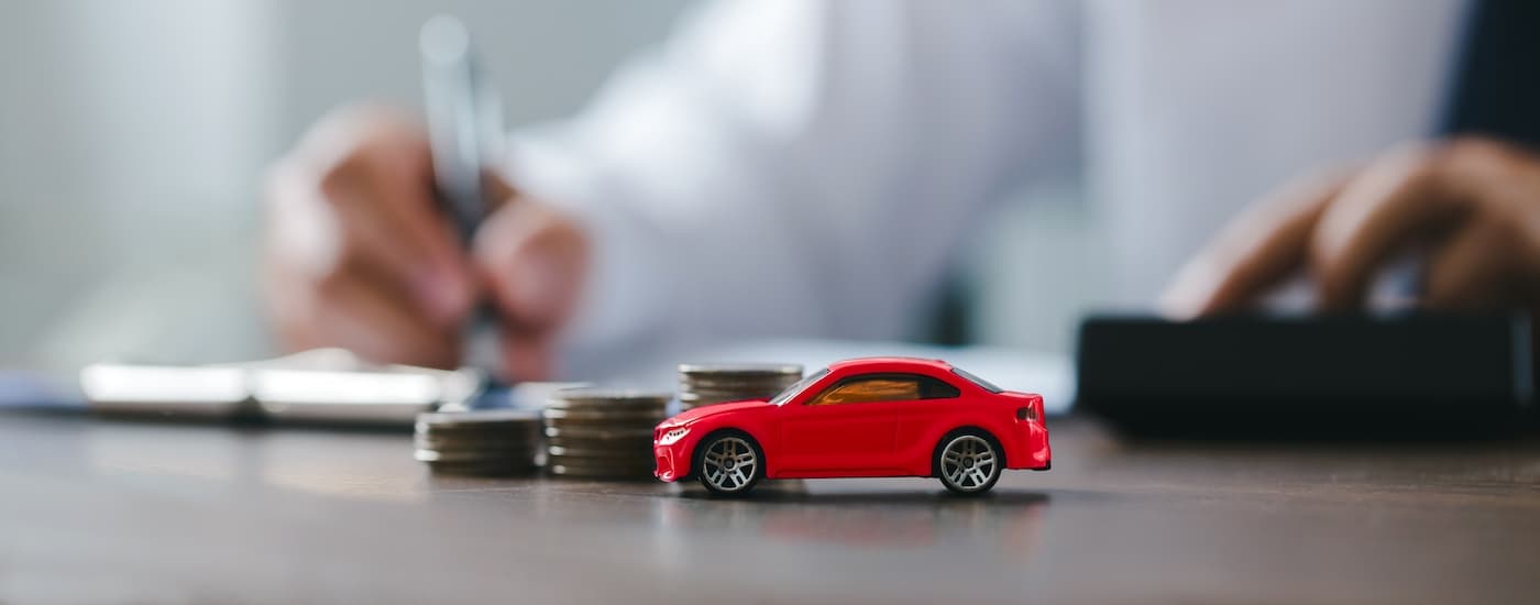 Red toy car on a desk in front of three stacks of coins and a man filling out paperwork