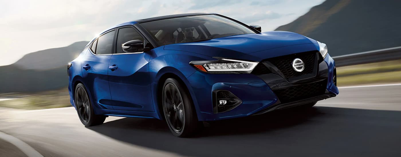 A blue 2021 Nissan Maxima is shown driving on an open road after visiting a used Nissan dealership.