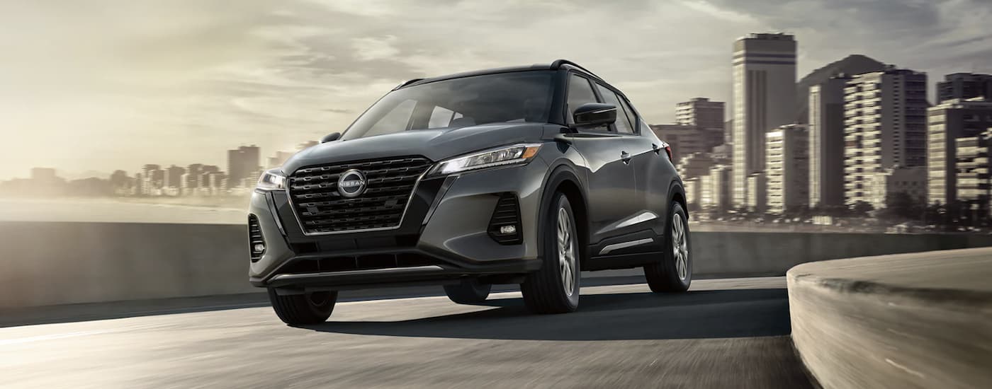 A grey 2022 Nissan Kicks is shown from the front driving on an open road past a city.