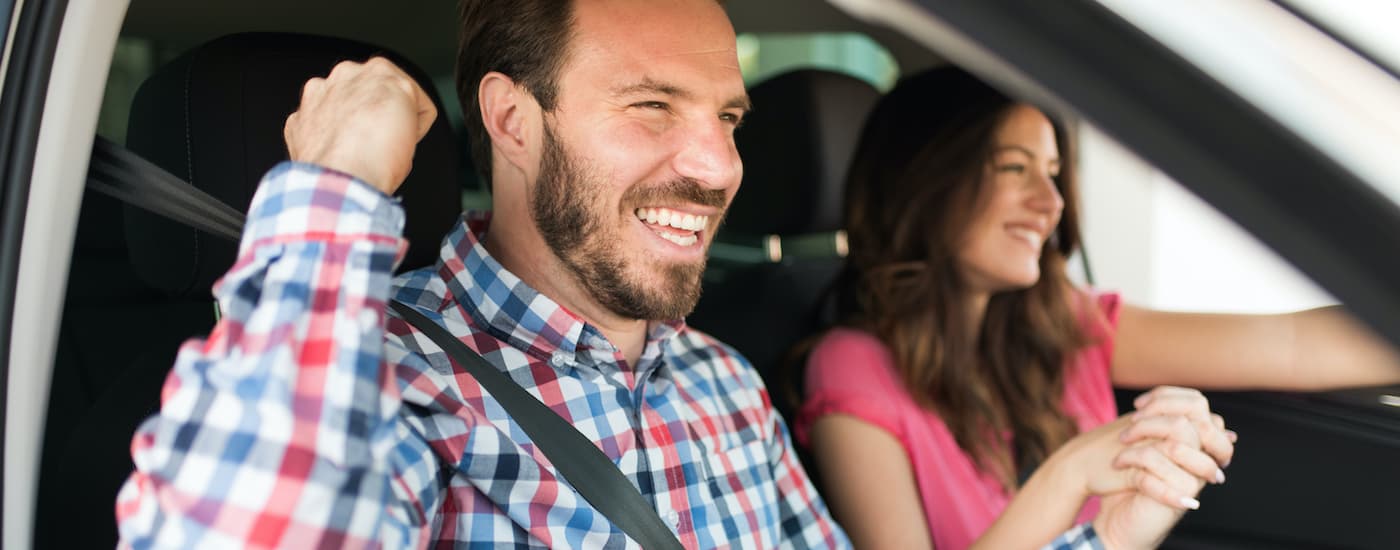 A couple is shown sitting in the front seat of a car while holding hands and smiling.
