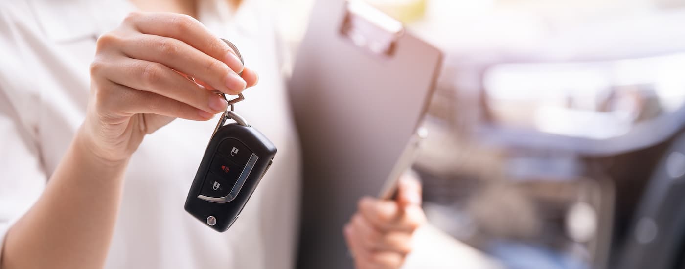 A saleswoman is shown holding a car key and clipboard at a car dealership.