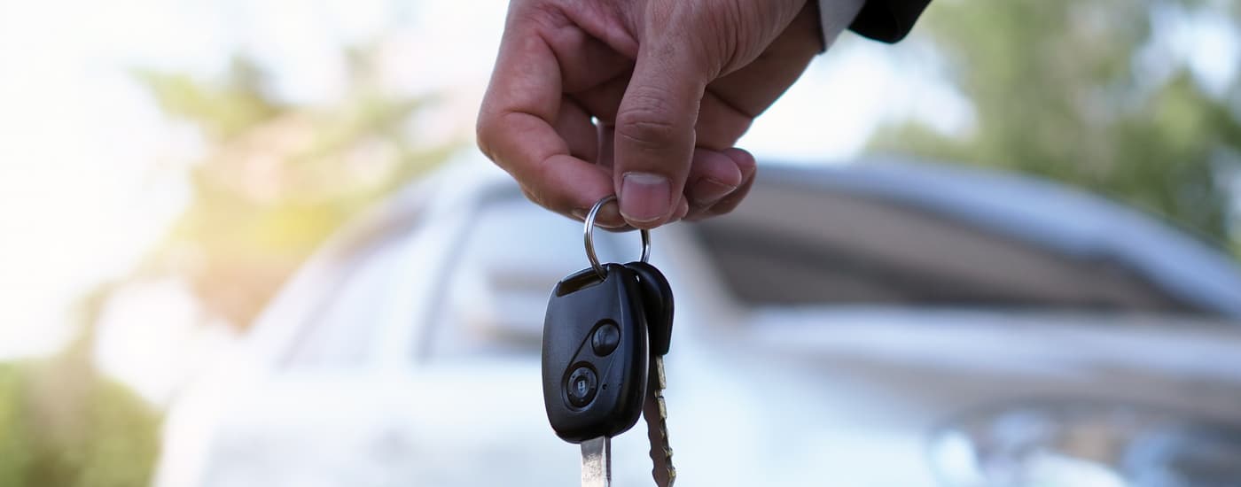 A hand is shown holding a set of car keys in front of a blurred silver vehicle.