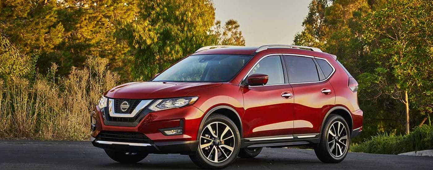 A red 2020 used Nissan Rogue for sale is shown parked in a tree-lined parking lot.