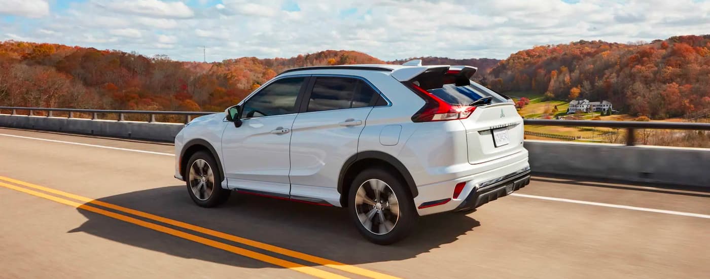 A white 2023 Mitsubishi Eclipse Cross is shown driving over a bridge past autumn leaves.