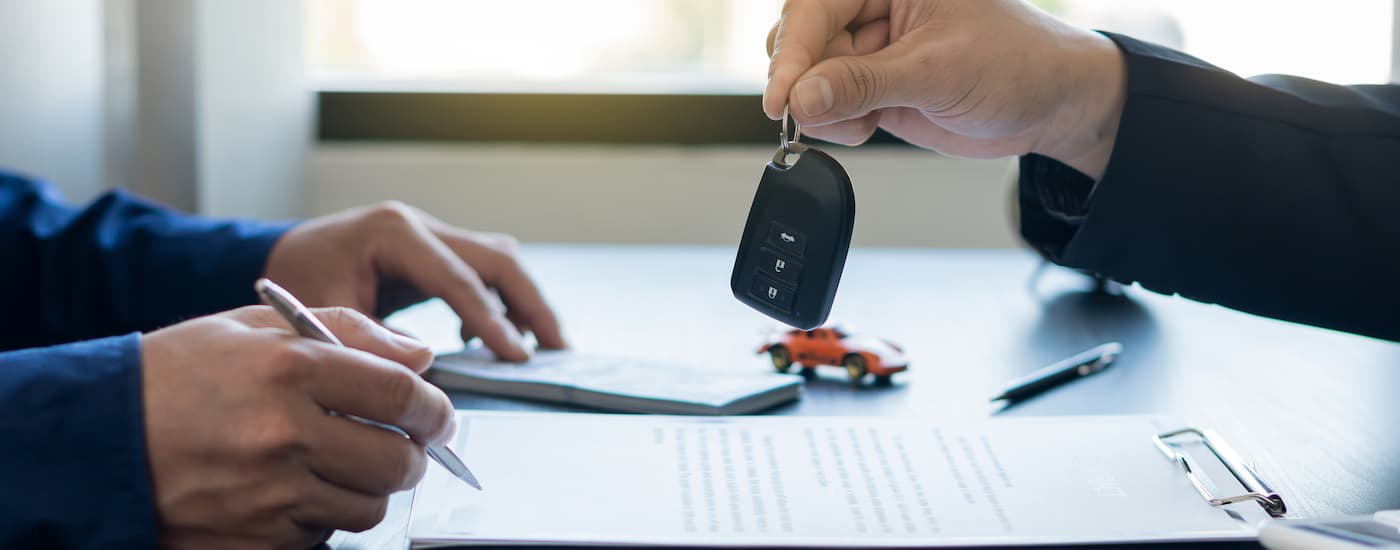 A car salesman is shown handing over a car key while a customer fills out paperwork.