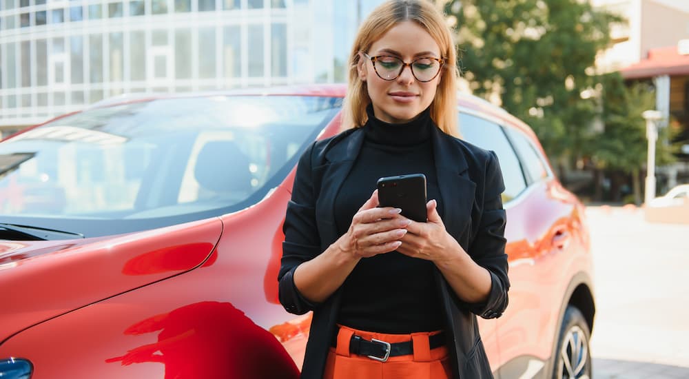 A woman is shown standing next to a red vehicle as she searches 'how to sell my car' on her phone.