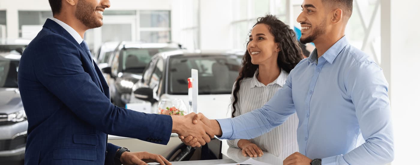 A salesman is shown shaking hands with a man inside of a car dealership.