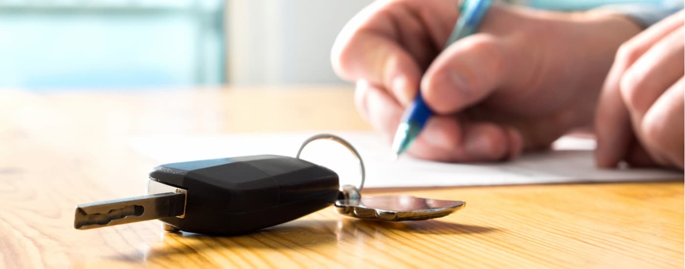 A close up of a car key next to a hand filing out paperwork on a wooden table is shown.