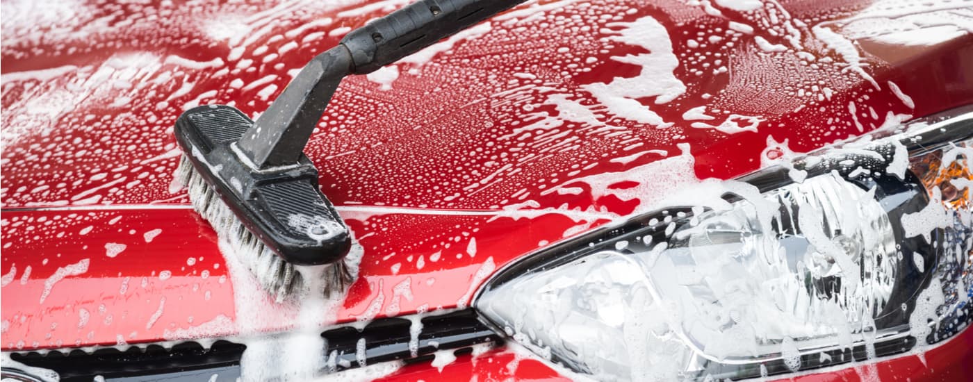 A brush is shown cleaning the soap-covered hood of a red sedan.