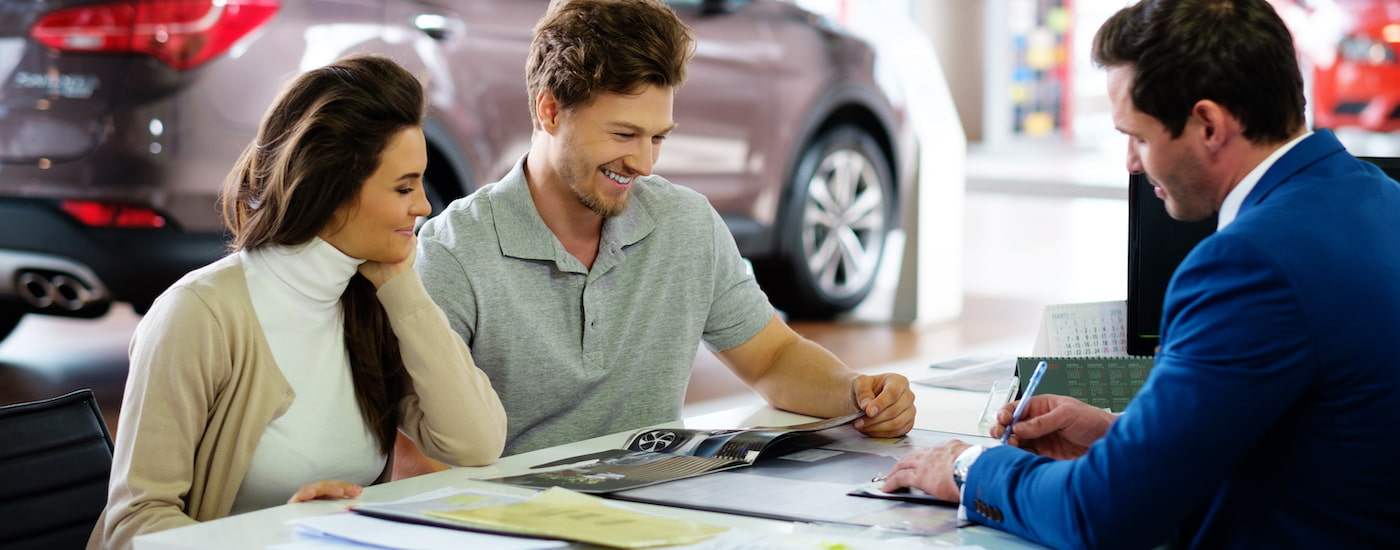 Two people are shown talking to a salesperson at a car dealership.