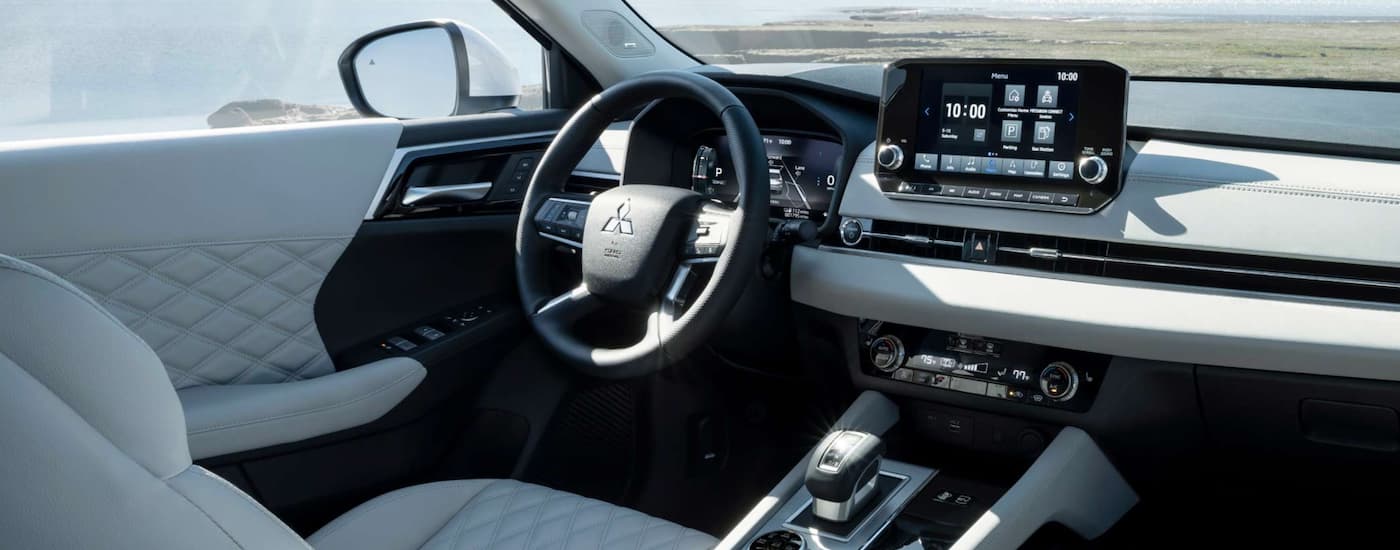 The white interior of a 2023 Mitsubishi Outlander PHEV shows the steering wheel and infotainment screen.