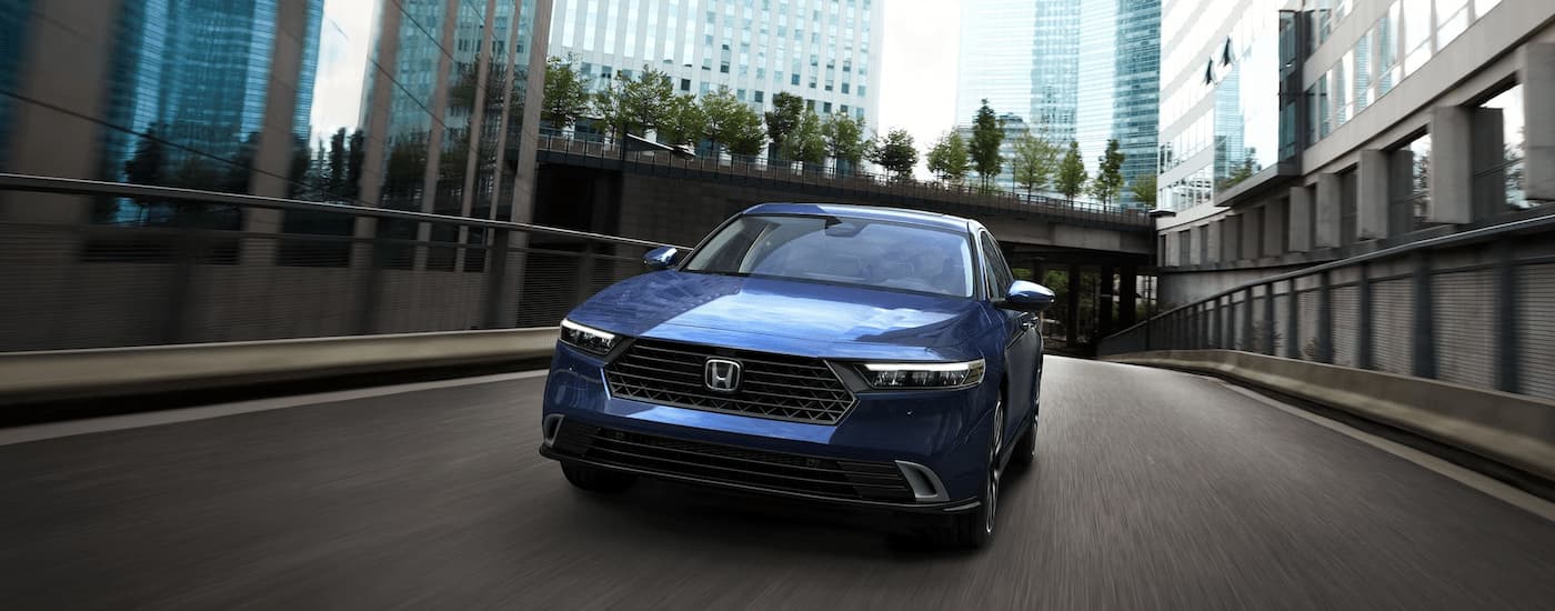 A blue 2023 Honda Accord Sport Hybrid is shown from the front driving on a city highway.