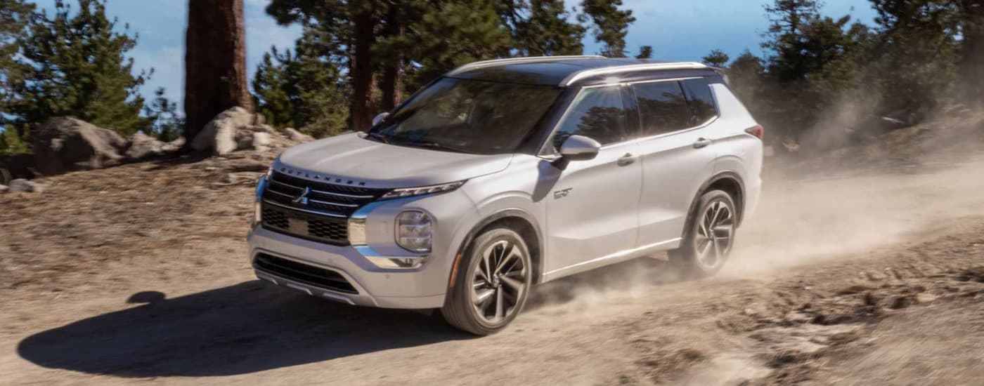 A white 2023 Mitsubishi Outlander PHEV is shown from the side driving on a dirt road.