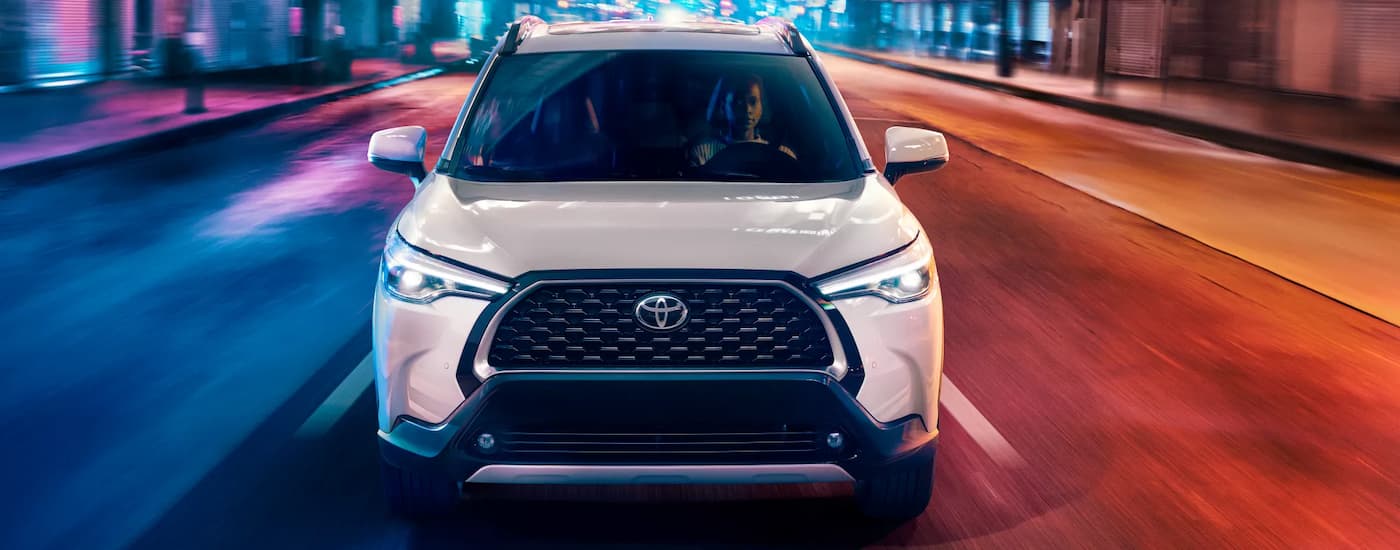 View from the front of a white 2022 Toyota Corolla Cross driving on a colorfully lit city street.
