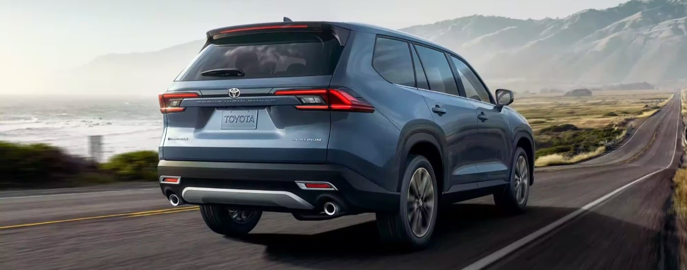 A blue 2024 Toyota Grand Highlander Platinum is shown from a rear angle driving on an open highway.