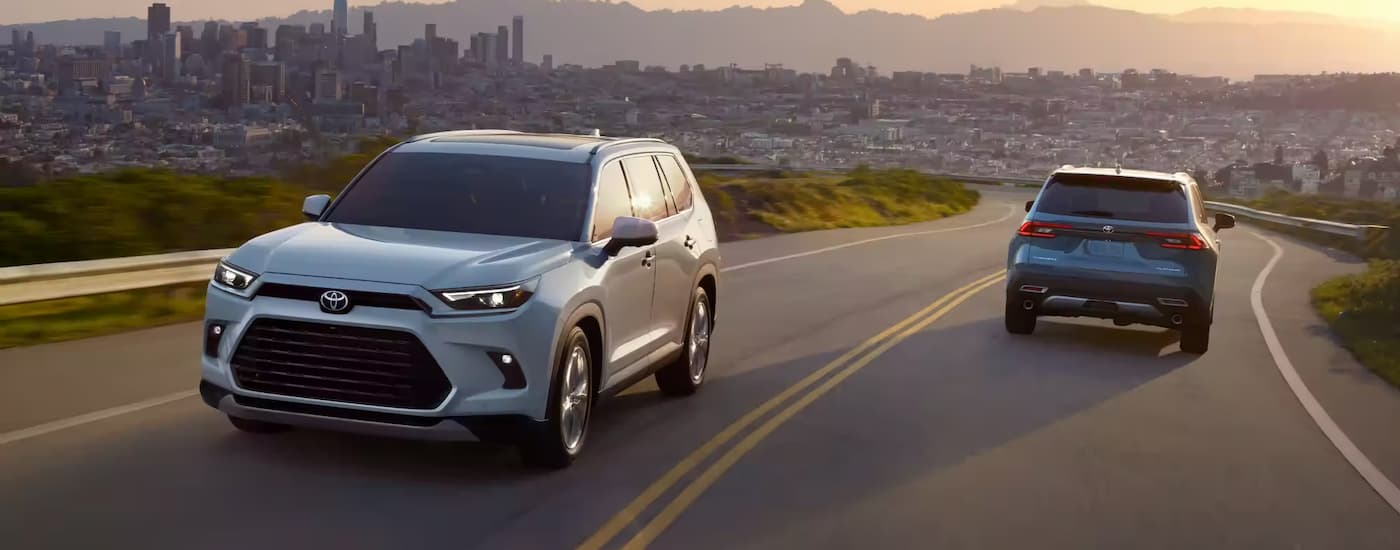 A white and a blue 2024 Toyota Grand Highlander are shown crossing paths on a winding road.