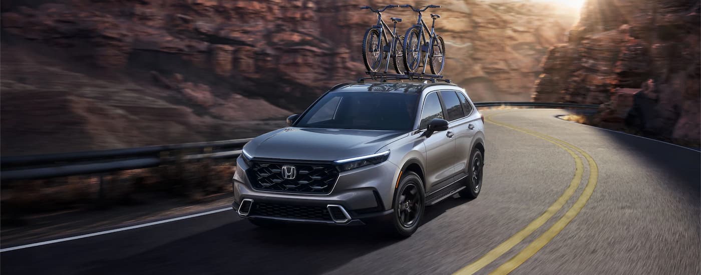 A silver 2023 Honda CR-V Hybrid with bicycles on the roof driving through a rocky canyon road.