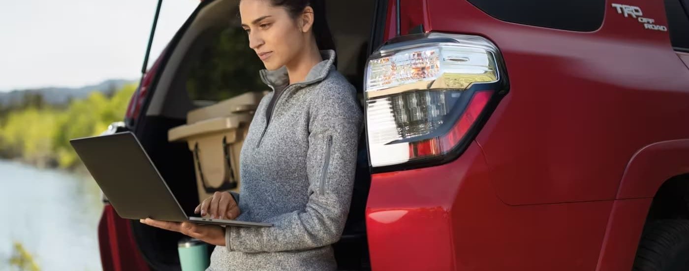 A person is shown leaning against a red 2023 Toyota 4Runner TRD Off-Road while using a laptop.