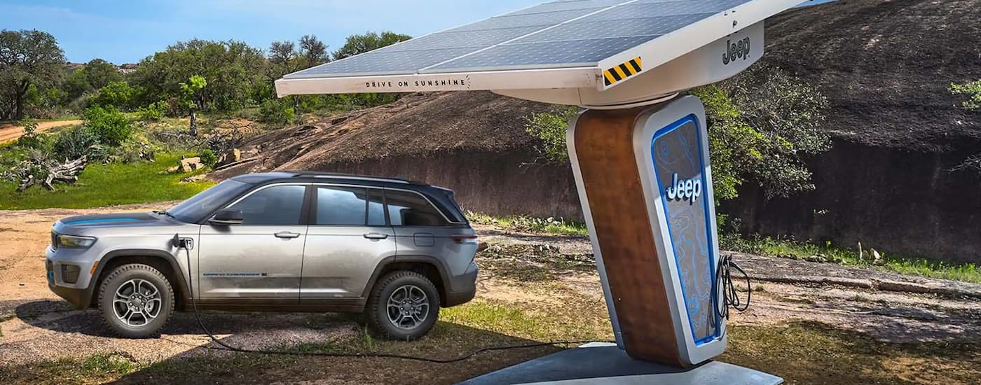 A silver 2023 Jeep Grand Cherokee 4xe is shown parked at a remote charging station near a trailhead.