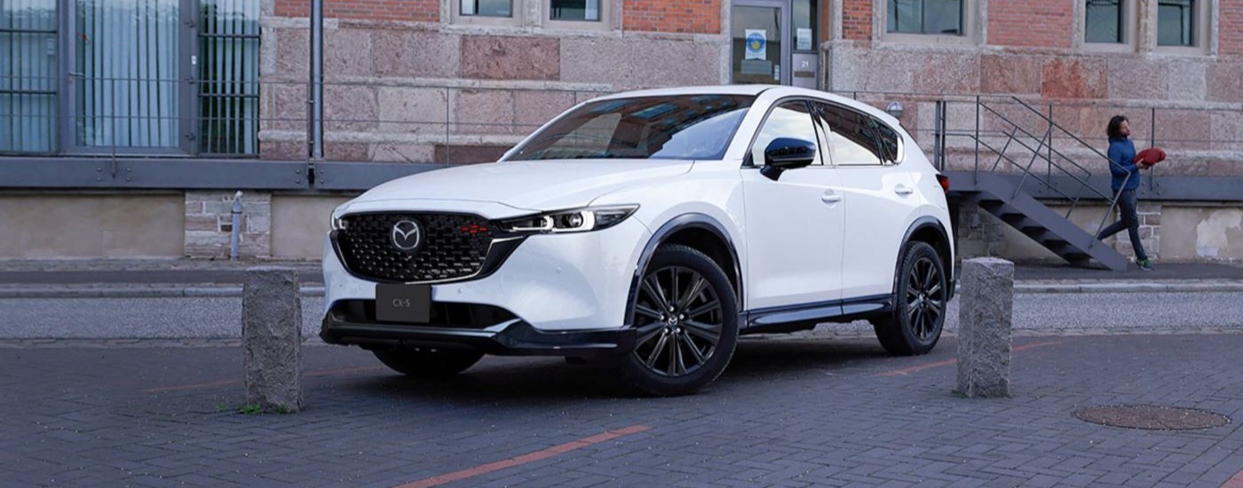 A white 2023 Mazda CX-5 is shown in front of a building after leaving a Kenmore Mazda dealer.