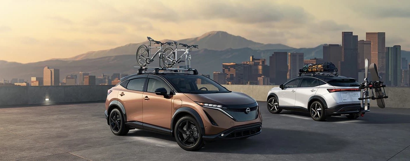A bronze and a white 2023 Nissan Ariya is shown near a city.
