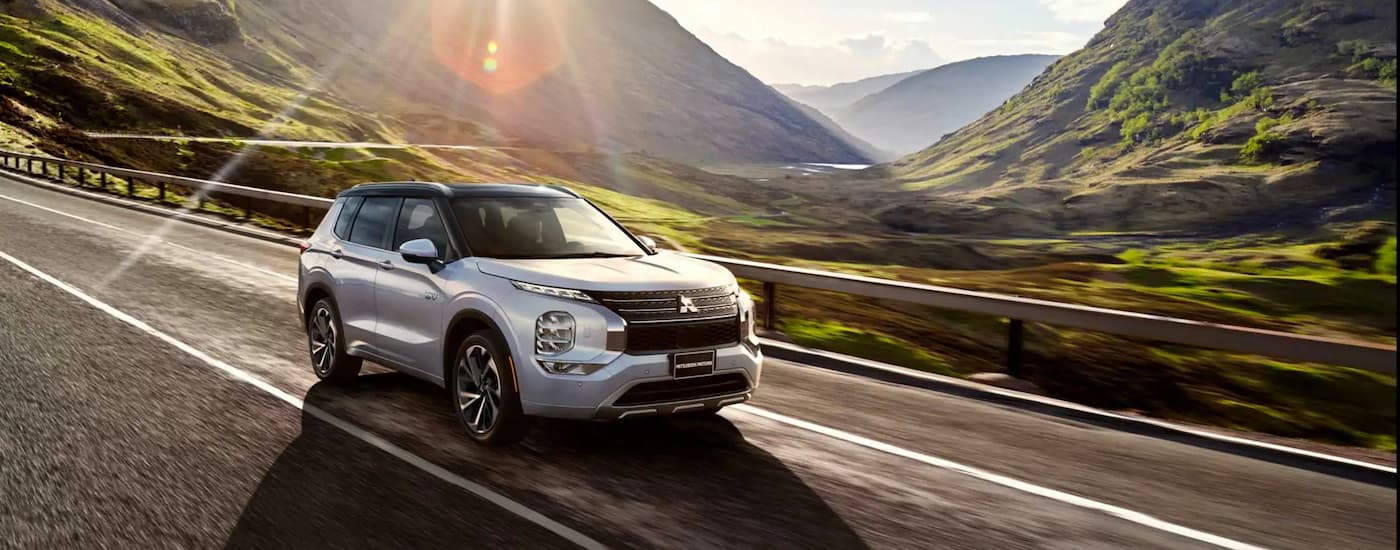 A silver 2023 Mitsubishi Outlander PHEV is shown driving on an open road through the mountains.
