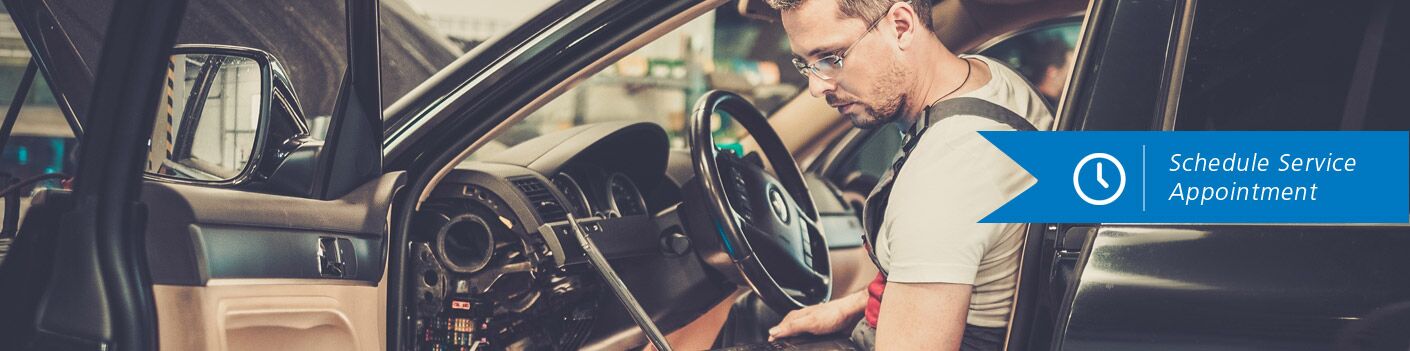 Man cradles a laptop and examines a vehicle's electrical system. Banner reads, "Schedule Service …