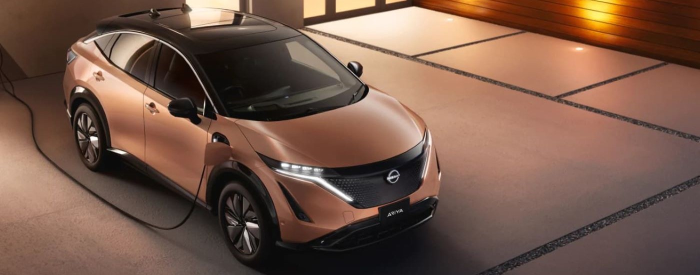 A bronze 2023 Nissan Ariya is shown charging on a driveway.