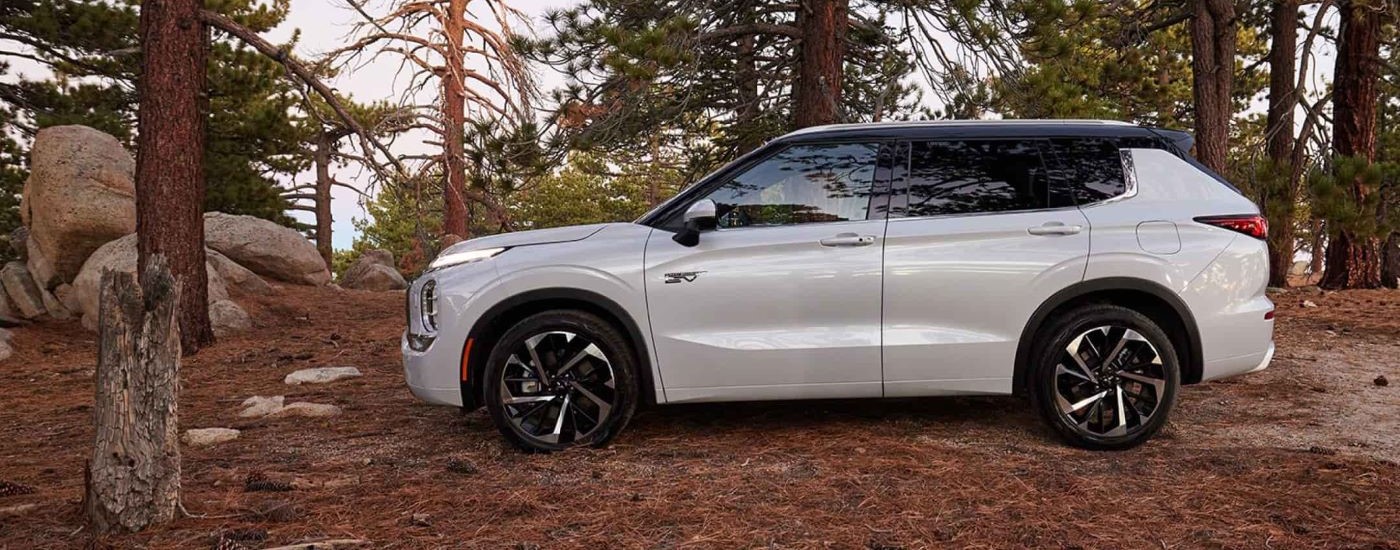 A white 2023 Mitsubishi Outlander PHEV is shown from the side near evergreen trees.