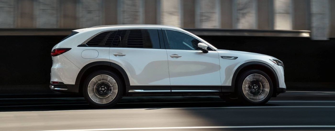A white 2024 Mazda CX-90 is shown from the side driving on a city street.