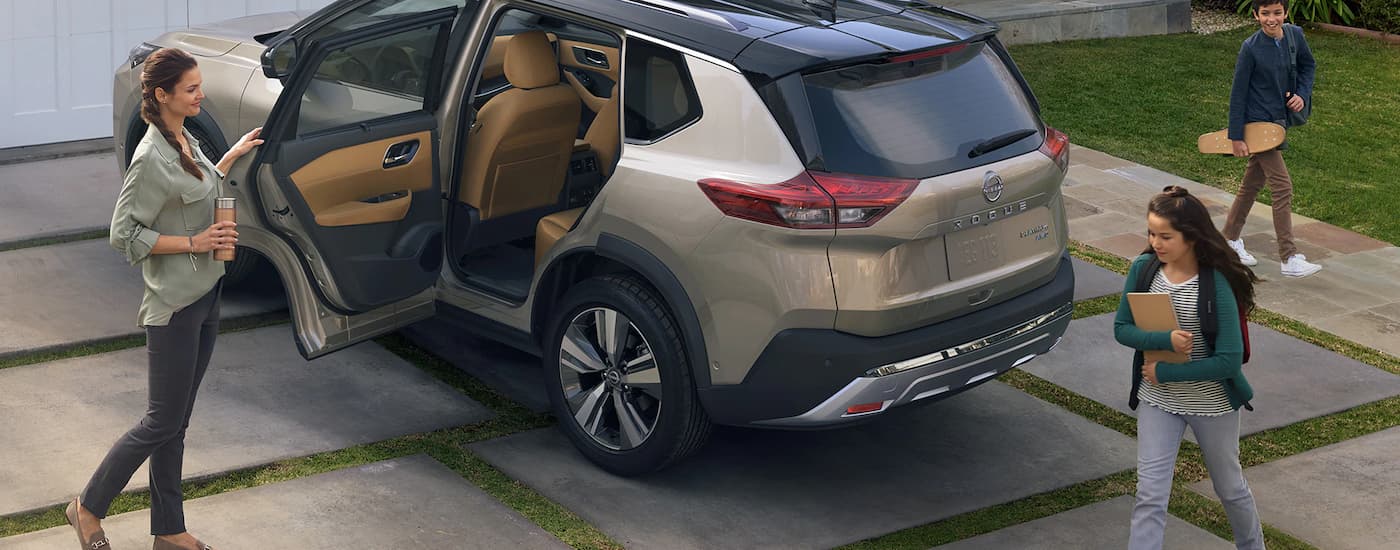 A family is shown walking around a tan 2023 Nissan Rogue Platinum parked on a driveway.