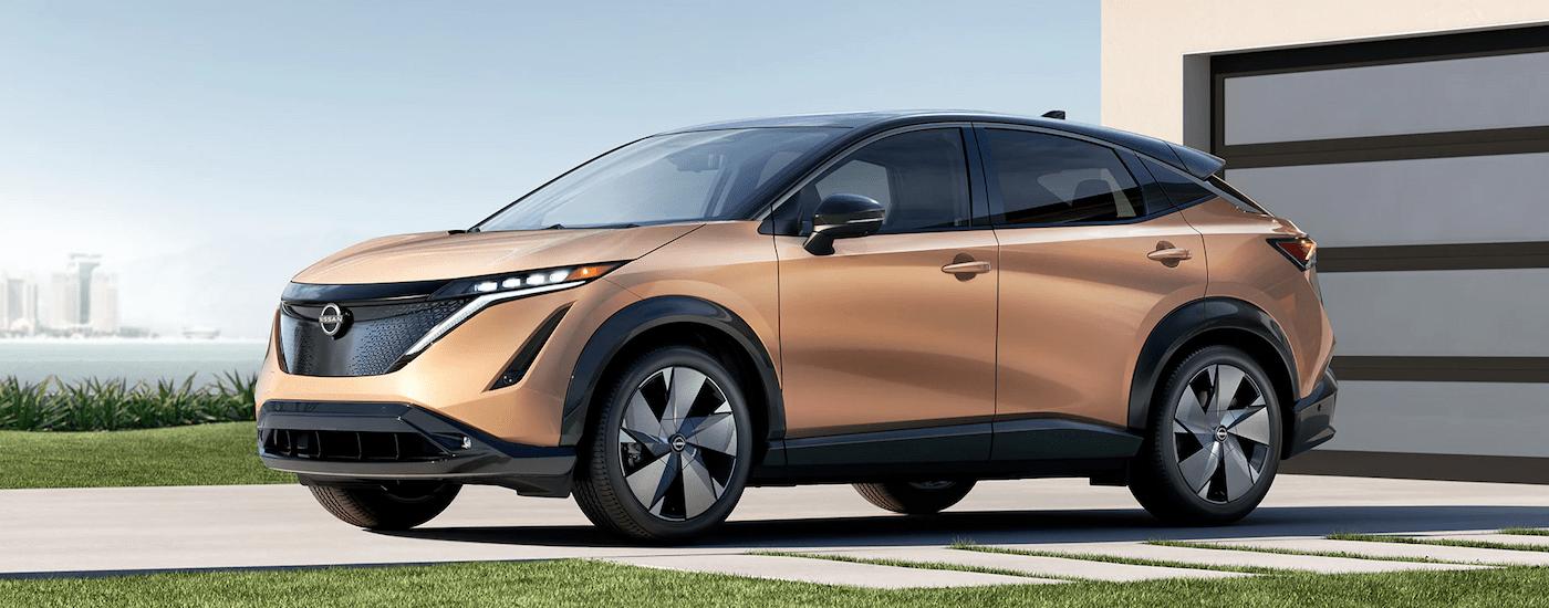 A tan 2023 Nissan Ariya is shown parked in a driveway in front of a garage.