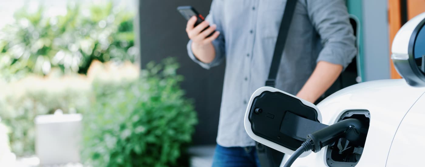 A man is shown standing next to vehicle charging while using his phone to search for EVs for Sale Near St. Albert.