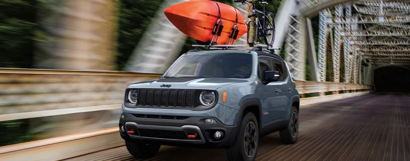 A grey 2023 Jeep Renegade is shown with an orange kayak on the roof while driving over a large bridge.