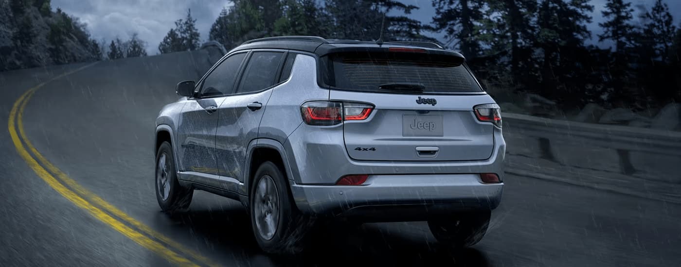 A silver 2023 Jeep Compass is shown from the rear driving on a winding road during a rainstorm.