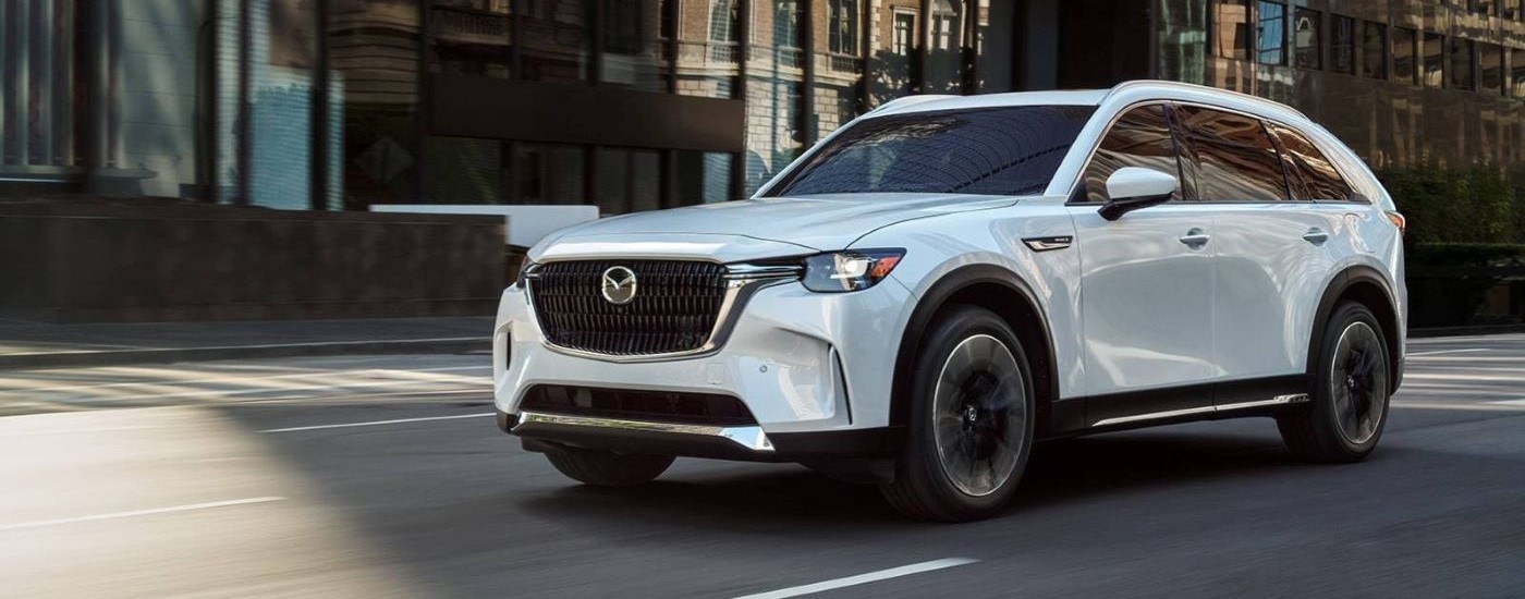 A white  2024 Mazda CX-90 PHEV is shown driving on a city street after visiting a Kirkland Mazda dealer.