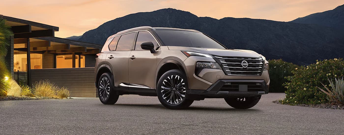 A tan 2024 Nissan Rogue for sale is shown parked in a driveway with a mountain view.