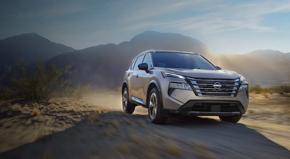 A grey 2024 Nissan Rogue for sale is shown driving on a dirt road.