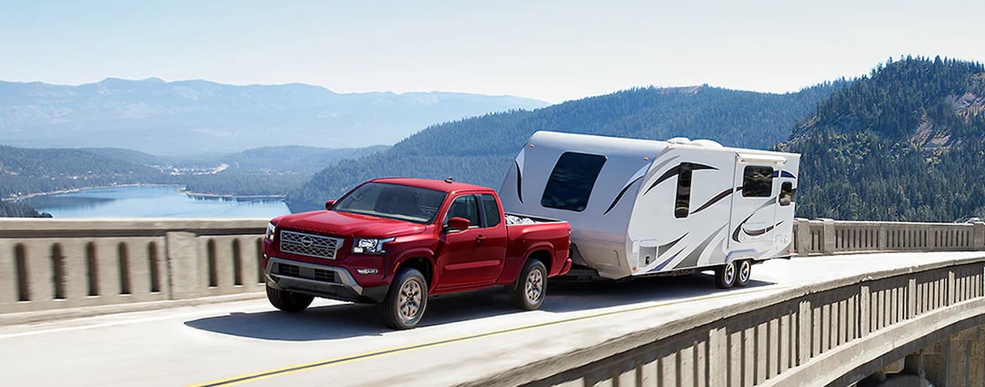 A red 2024 Nissan Frontier King Cab is shown towing a white camper trailer on a bridge.