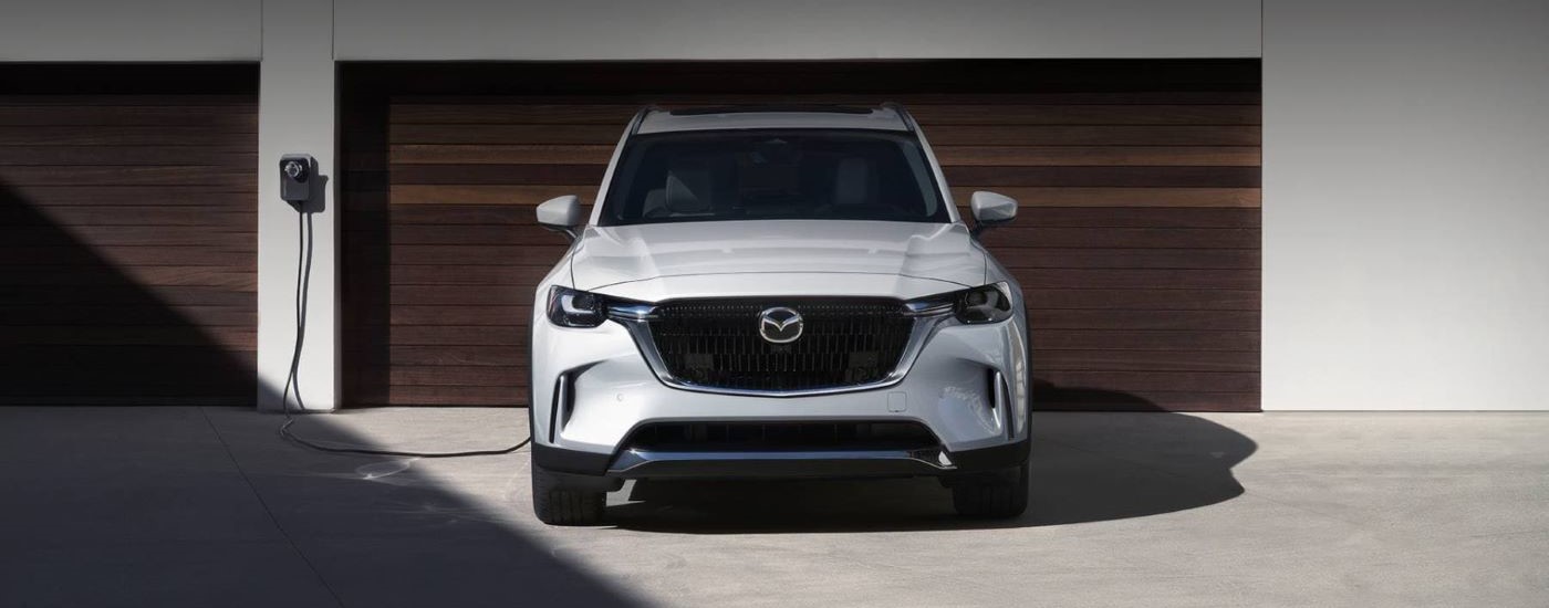 A white 2024 Mazda CX-90 PHEV is shown charging in front of a garage.
