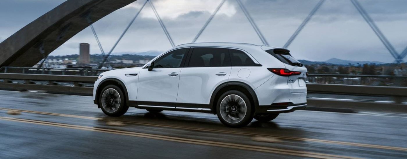 A white 2024 Mazda CX-90 PHEV is shown driving over a bridge after leaving a Mazda CX-90 dealer.