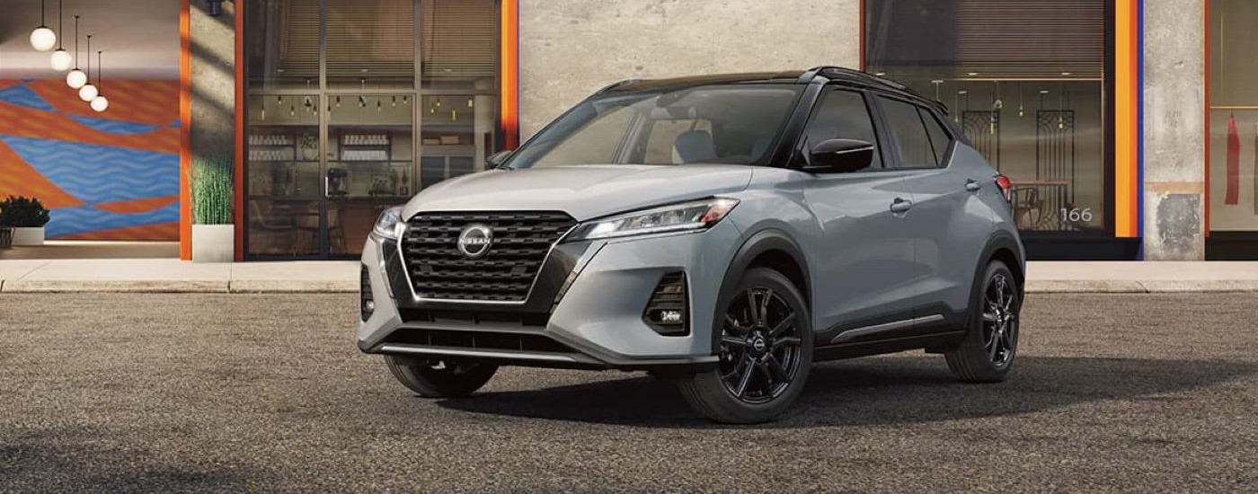 A grey 2024 Nissan Kicks for sale is shown parked in front of a shop.
