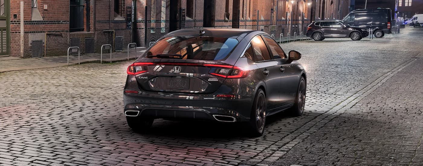 A grey 2024 Honda Civic Hatchback Sport Touring is shown from a rear angle driving on a stone street.
