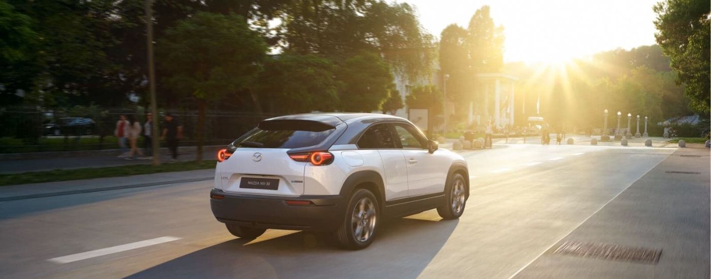 A white 2023 Mazda MX-30 is shown driving on a city street on a sunny day.