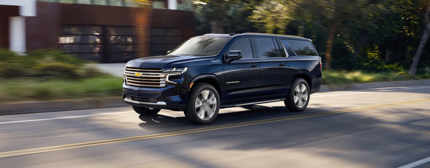 A dark blue 2021 Chevy Suburban High Country, a popular used SUV for sale, driving on a tree-lined road.
