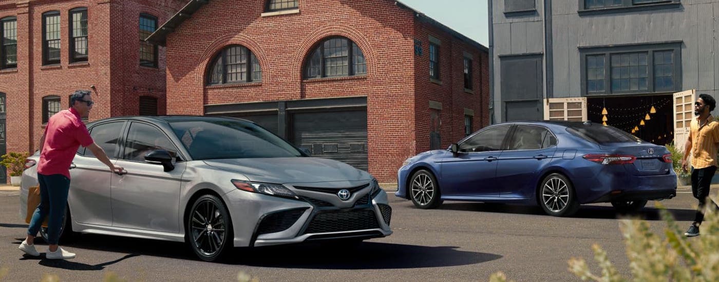 A grey and a blue 2024 Toyota Camry are parked in a city parking lot.