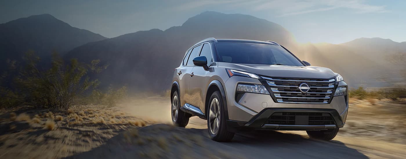 A grey 2024 Nissan Rogue is driving on a dusty dirt road after viewing cars for sale.